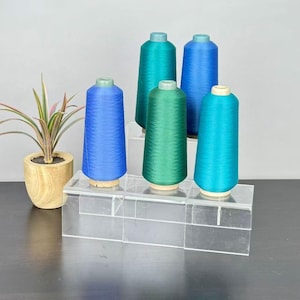 Peut inclure: Cinq bobines de fil coniques dans des tons de bleu et de vert, disposées sur un présentoir en acrylique transparent à plusieurs niveaux. Les couleurs incluent le bleu roi, le sarcelle, le vert forêt et le turquoise. Une plante est à gauche.