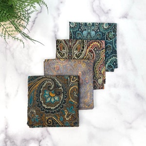 May include: Five folded paisley pocket squares in various colors, including teal, gold, and gray. The fabric has an ornate, swirling pattern. The pocket squares are arranged in a staggered, layered display.