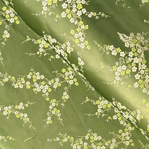 Peut inclure: Tissu vert avec un motif floral de fleurs blanches et jaunes sur des branches. Le tissu a un éclat subtil, et le motif se répète sur la surface. Les fleurs sont regroupées, créant une esthétique délicate et naturelle.