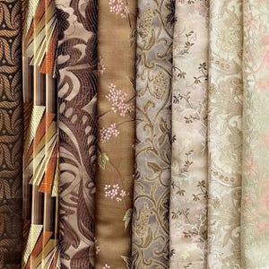 Op de afbeelding: Een verzameling verschillende stofstalen met ingewikkelde patronen en texturen. De stoffen tonen een reeks kleuren, waaronder bruin, goud en crème, met bloemen- en geometrische ontwerpen. Sommige stalen hebben geborduurde bloemendetails.