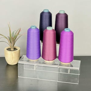 May include: Five conical spools of thread in shades of purple, magenta, and dark violet are arranged on a clear acrylic tiered display stand. Each spool features a light blue top cap. A small potted plant with variegated leaves sits nearby.