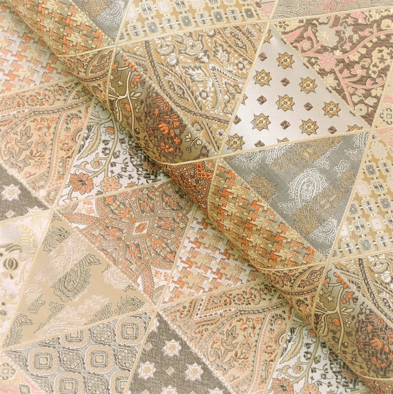 Op de afbeelding: Opgerolde stof met een patchwork-ontwerp. De stof heeft geometrische vormen, waaronder driehoeken en vierkanten, in beige, bruine en oranje tinten. Ingewikkelde paisley- en bloempatronen sieren de stof en cre&euml;ren een textuur en gedetailleerd uiterlijk.
