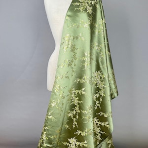 May include: A bolt of olive green fabric draped over a mannequin. The fabric is a satin with a floral pattern in white and gold. The flowers are small and clustered, creating a delicate design.