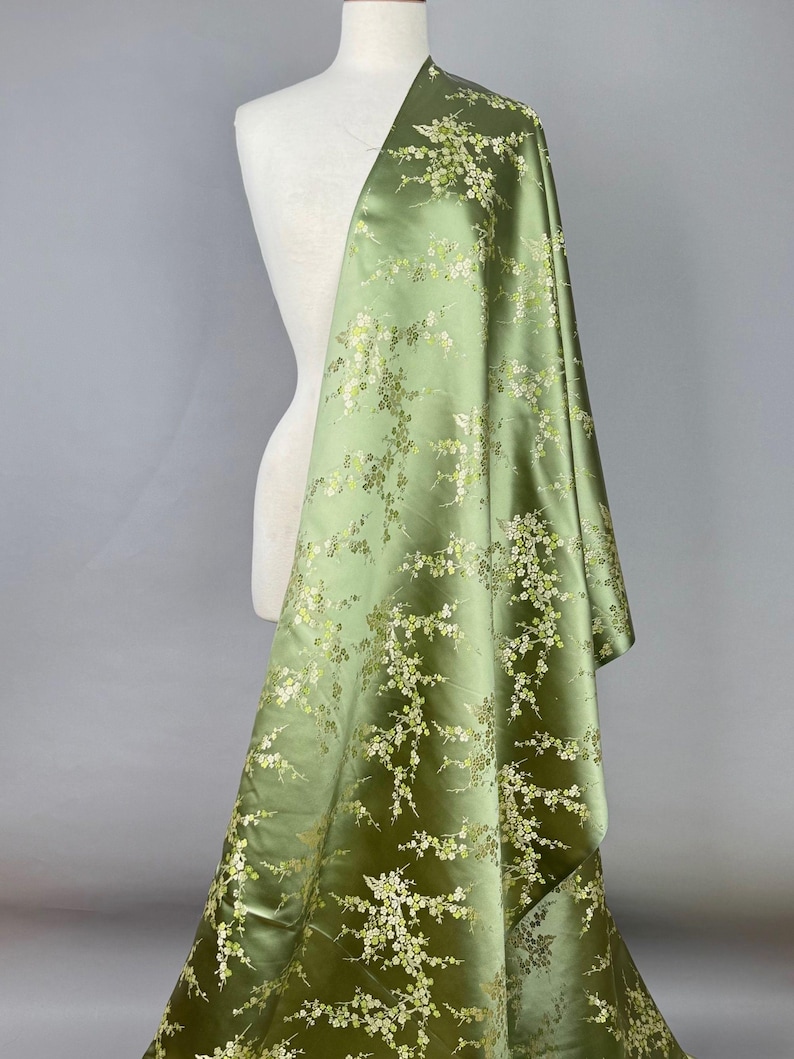 May include: A bolt of olive green fabric draped over a mannequin. The fabric is a satin with a floral pattern in white and gold. The flowers are small and clustered, creating a delicate design.