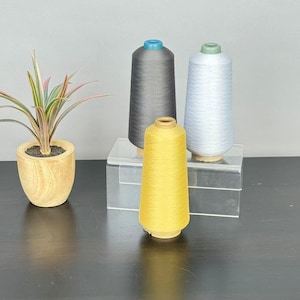 May include: Three conical spools of thread in dark gray, light blue, and mustard yellow, each featuring a small plastic cap at the top. The spools are arranged on a clear acrylic stand next to a small potted plant with variegated leaves.