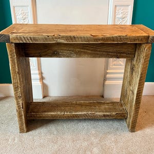Pode incluir: Uma consola retangular de madeira rústica com uma prateleira inferior. A mesa é construída com madeira recuperada, mostrando grãos e nós naturais. As pernas são anguladas. A mesa tem aproximadamente 91 cm de comprimento, 30 cm de largura e 76 cm de altura.