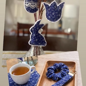 Może przedstawiać: Asortyment dekoracji i akcesoriów w tematyce wielkanocnej. Obejmuje niebieskie, materiałowe ozdoby w kształcie jajka, tulipana i królika z białymi wzorami kwiatowymi. Prezentowane są również filiżanka herbaty, podstawka, gumka do włosów i brelok.