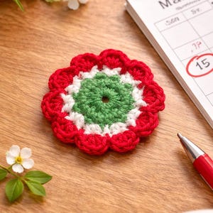 Könnte beinhalten: Ein handgefertigter gehäkelter Blumenuntersetzer in Rot, Weiß und Grün liegt auf einer Holzoberfläche. Im Hintergrund befinden sich ein Kalender mit dem 15. März, ein roter Stift und Blumen. Der Untersetzer hat einen gewellten Rand.