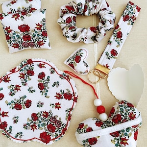 Könnte beinhalten: Eine Auswahl an weißen Stoffaccessoires mit einem roten Blumenmuster. Die Artikel umfassen zwei kleine Kissen, ein Haargummi, eine Handschlaufe, einen herzförmigen Schlüsselanhänger und einen Untersetzer. Sie sind auf einer cremefarbenen Oberfläche angeordnet.