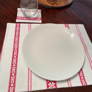 May include: A white rectangular placemat featuring red embroidered geometric patterns and stripes, topped with a plain white round dinner plate. A matching square coaster sits nearby, supporting a clear glass with etched text on its surface.