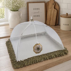 Op de afbeelding: Een witte voedselafdekking van gaas met een decoratieve goud-zwarte rand. De afdekking heeft een bijen-thema bedel die van bovenaf hangt. Het aanrecht heeft ook een witte vaas met bloemen, een bord met "Home Sweet Home" en houten snijplanken.