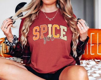 May include: A maroon long-sleeve t-shirt with the text "Spice girl" in a retro-style font with orange, yellow, and white accents.
