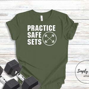 May include: Olive green t-shirt with white text that reads "Practice Safe Sets" and a white graphic of crossed dumbbells inside a circle.  The t-shirt is on a wood background with two black dumbbells.