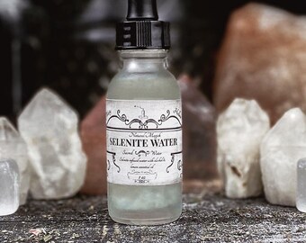 May include: A clear glass bottle with a dropper top, filled with a light green liquid. The bottle has a white label with black text that reads "Natural Magick Selenite Water Sacred Water Selenite-infused water with alcohol & lemon essential oil 1 fl oz F-269". The bottle is sitting on a wooden surface with white crystals around it.