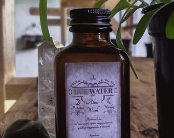 May include: A brown glass bottle labeled "Rune Water" with text for altar use, protection, wash, and warding. The bottle is next to green stones and a clear crystal. The bottle contains 2 fl oz of rune-infused water.