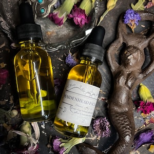 Puede incluir: Dos botellas de vidrio &aacute;mbar con goteros, llenas de un l&iacute;quido dorado. Las botellas est&aacute;n etiquetadas con "Maidenhead Facial" y una lista de ingredientes bot&aacute;nicos. Las botellas est&aacute;n rodeadas de flores secas y una figura de sirena de metal.