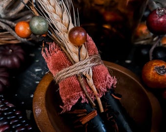 May include: A rustic arrangement featuring a wooden bowl holding a bundle of dried wheat, cinnamon sticks, and a small piece of red fabric tied with twine. Decorative berries and a dark glass bottle are in the background.