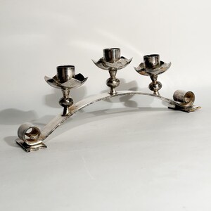 Può includere: Candelabro d'argento con tre portacandele. Il candelabro ha un design curvo con volute decorative a ciascuna estremità. I portacandele hanno una forma a fiore.