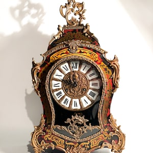 May include: A gold-toned mantle clock with ornate details and a black face with Roman numerals. The clock has a red and gold painted design.