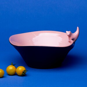 May include: A pink and black ceramic bowl with a stylised animal handle. Three small yellow tomatoes are in front of the bowl on a blue background.