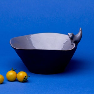 May include: A grey ceramic bowl with a cat-shaped handle. The bowl is on a blue surface with three yellow cherry tomatoes in front of it.