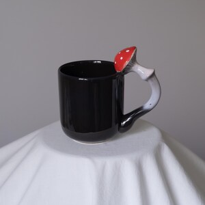 May include: Black ceramic mug with a red and white mushroom handle. The mushroom is shaped like a toadstool with white spots.