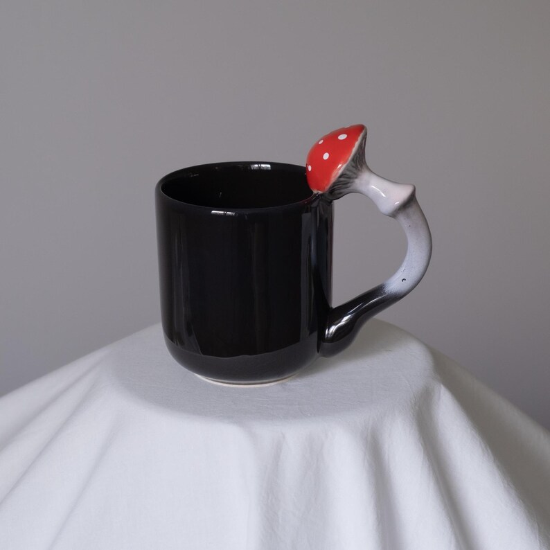 May include: Black ceramic mug with a red and white mushroom handle. The mushroom is shaped like a toadstool with white spots.