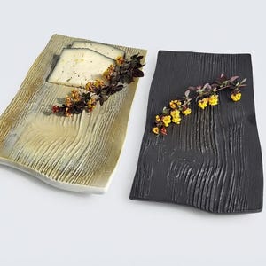 May include: Two rectangular serving platters with a textured wood grain surface. One platter is finished in a metallic gold tone and holds two slices of cheese garnished with a sprig of small yellow flowers. The other platter is matte black.