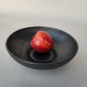 Puede incluir: Un cuenco de cerámica negro con una manzana roja en su interior. El cuenco tiene un diseño ancho y poco profundo con un acabado mate oscuro. La manzana es de un rojo vibrante con vetas amarillas.