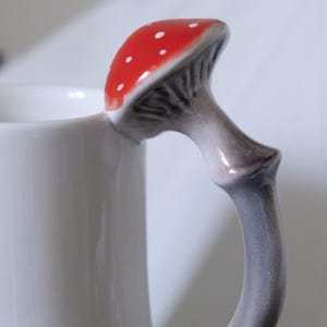 May include: White ceramic mug with a mushroom-shaped handle. The mushroom is red with white dots and a gray stem.