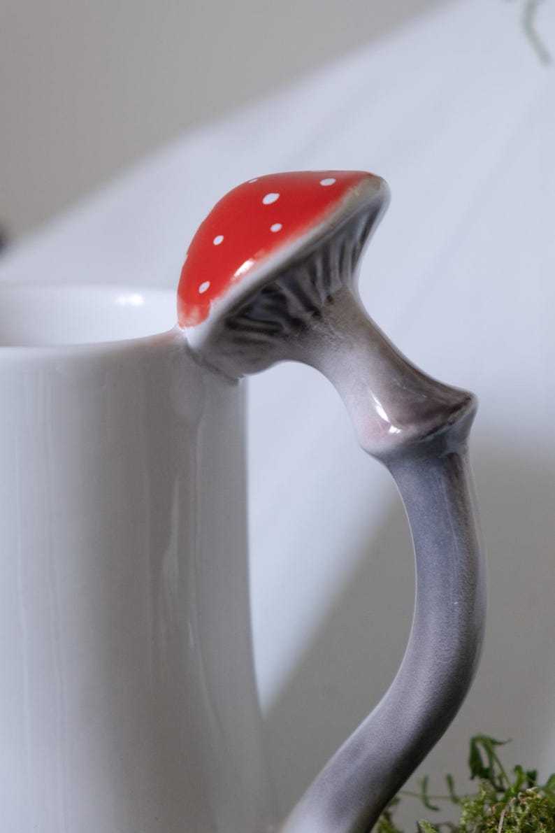May include: White ceramic mug with a mushroom-shaped handle. The mushroom is red with white dots and a gray stem.