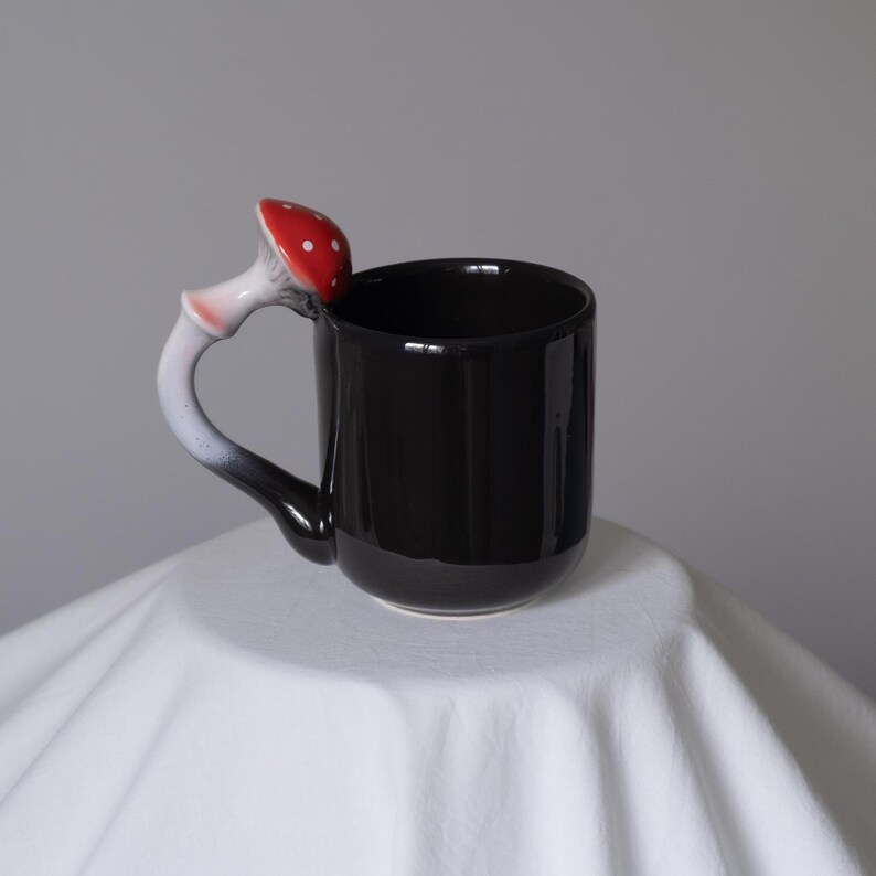 May include: Black ceramic mug with a red and white mushroom shaped handle. The mug is sitting on a white surface.