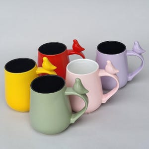Może przedstawiać: Kolekcja ceramicznych kubków w różnych kolorach: żółtym, czerwonym, zielonym, różowym i lawendowym. Każdy kubek ma uchwyt w kształcie ptaka i czarne wnętrze. Kubki są ustawione na jasnoszarej powierzchni.