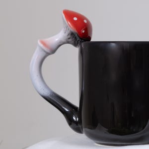 May include: Black ceramic mug with a white and red mushroom shaped handle. The mushroom has white dots on the red cap.