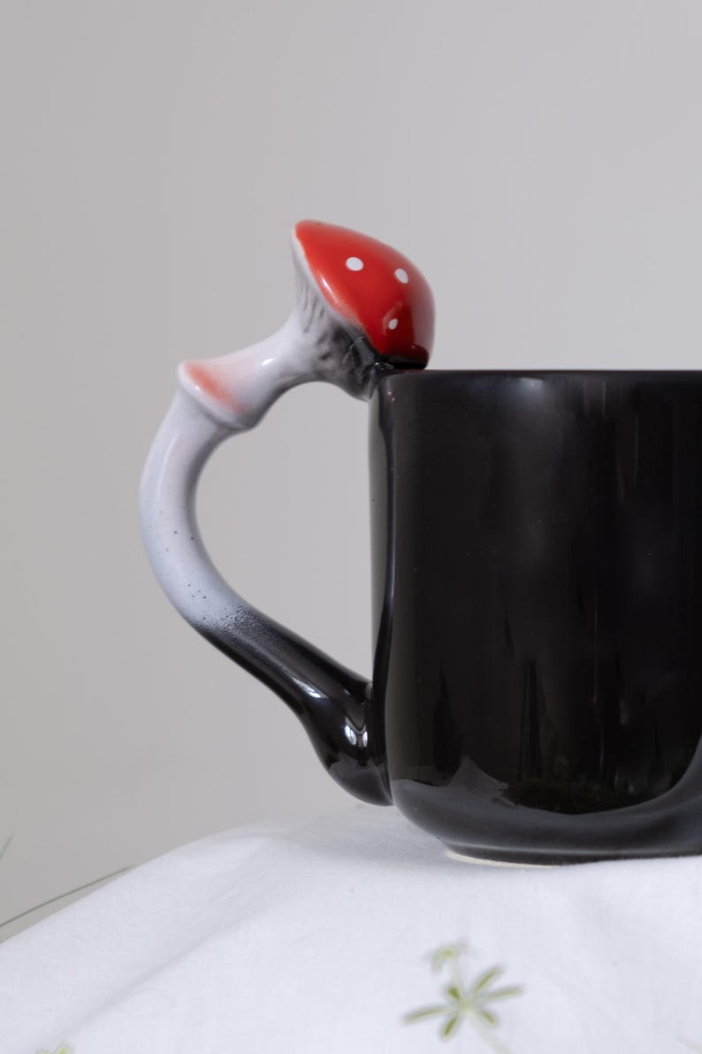 May include: Black ceramic mug with a white and red mushroom shaped handle. The mushroom has white dots on the red cap.