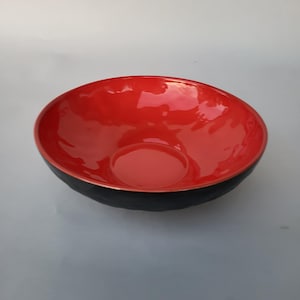 Może przedstawiać: Błyszcząca czerwona miska z czarnym zewnętrzem. Misa ma gładką, odblaskową powierzchnię i prosty design. Wnętrze jest żywo czerwone, kontrastujące z czarną krawędzią.