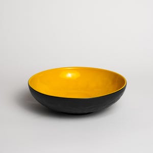 Op de afbeelding: Een ronde kom met een gladde, felgele binnenkant en een getextureerde, matzwarte buitenkant. De kom heeft een breed, open ontwerp met een ondiepe vorm, wat een duidelijk contrast laat zien tussen het heldergele binnenoppervlak en de donkere buitenafwerking.