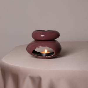 May include: A mauve ceramic oil diffuser with a round top and a smaller round bottom. The diffuser has a small opening in the bottom for a tea light candle. A lit tea light candle is inside the diffuser.