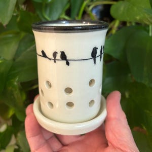 May include: A white ceramic oil warmer with a black rim and a series of small holes. The warmer features a black line with several black bird silhouettes perched on it. The warmer sits on a matching saucer.