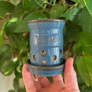 May include: A blue ceramic plant pot with a matching saucer. The pot has circular openings around the middle and decorative detailing. The rim and detailing have a brown glaze. The pot is held in front of green leaves. The pot is approximately 10 cm tall.