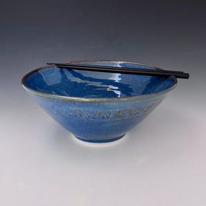 Può includere: Una ciotola di ceramica blu fatta a mano con una superficie strutturata e un bordo scuro. Bacchette nere poggiano sulla ciotola. La ciotola è di un blu intenso, con riflessi blu chiaro e bianchi.