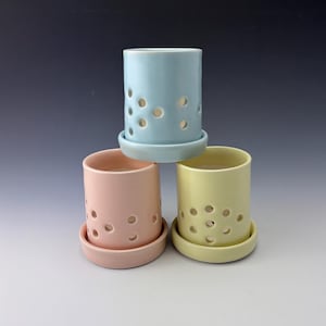 May include: Three ceramic candle holders in pastel colors: light blue, pink, and yellow. Each cylindrical holder has a matching saucer base and is perforated with several circular holes. The holders are stacked, showcasing their design.