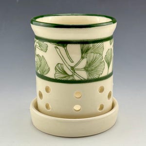 May include: A white ceramic planter with a green ginkgo leaf design and drainage holes. The planter has a matching saucer.