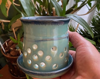 May include: A teal blue ceramic planter with drainage holes and a matching saucer. The planter is hand-crafted and has a glazed finish.