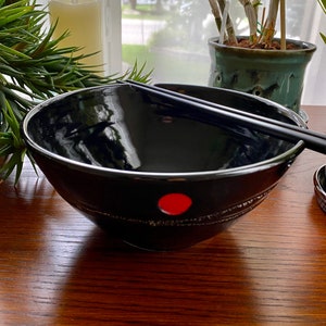 May include: A black ceramic bowl with a red dot and a silver rim. The bowl is on a wooden table with chopsticks resting on the rim.