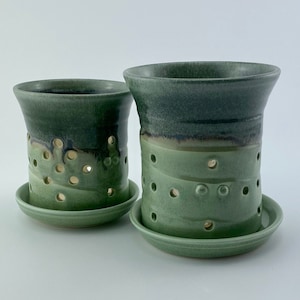 May include: Two handmade ceramic candle holders in shades of green. Each holder has a cylindrical shape with a flared top and a series of circular cutouts. They sit on matching round saucers. The glaze transitions from dark to light green.