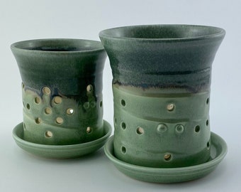May include: Two handmade ceramic candle holders in shades of green. Each holder has a cylindrical shape with a flared top and a series of circular cutouts. They sit on matching round saucers. The glaze transitions from dark to light green.