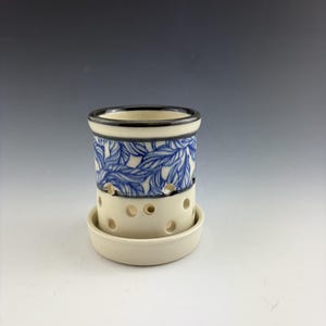 May include: A cylindrical ceramic vessel with a cream-colored base and a blue leaf pattern around the upper rim. The body features multiple circular cutouts and sits on a matching circular ceramic saucer with a dark rim.