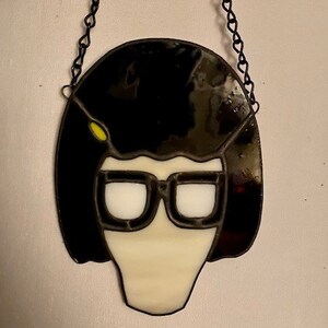 May include: A stained glass window hanging ornament featuring a cartoon character with black hair, white glasses, and a white face.