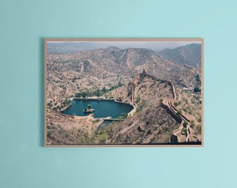 May include: A framed print of a scenic landscape featuring a lake surrounded by mountains and a fortress wall. The image has a warm color palette with shades of brown, green, and blue. The print is displayed on a light blue wall above a white sofa.